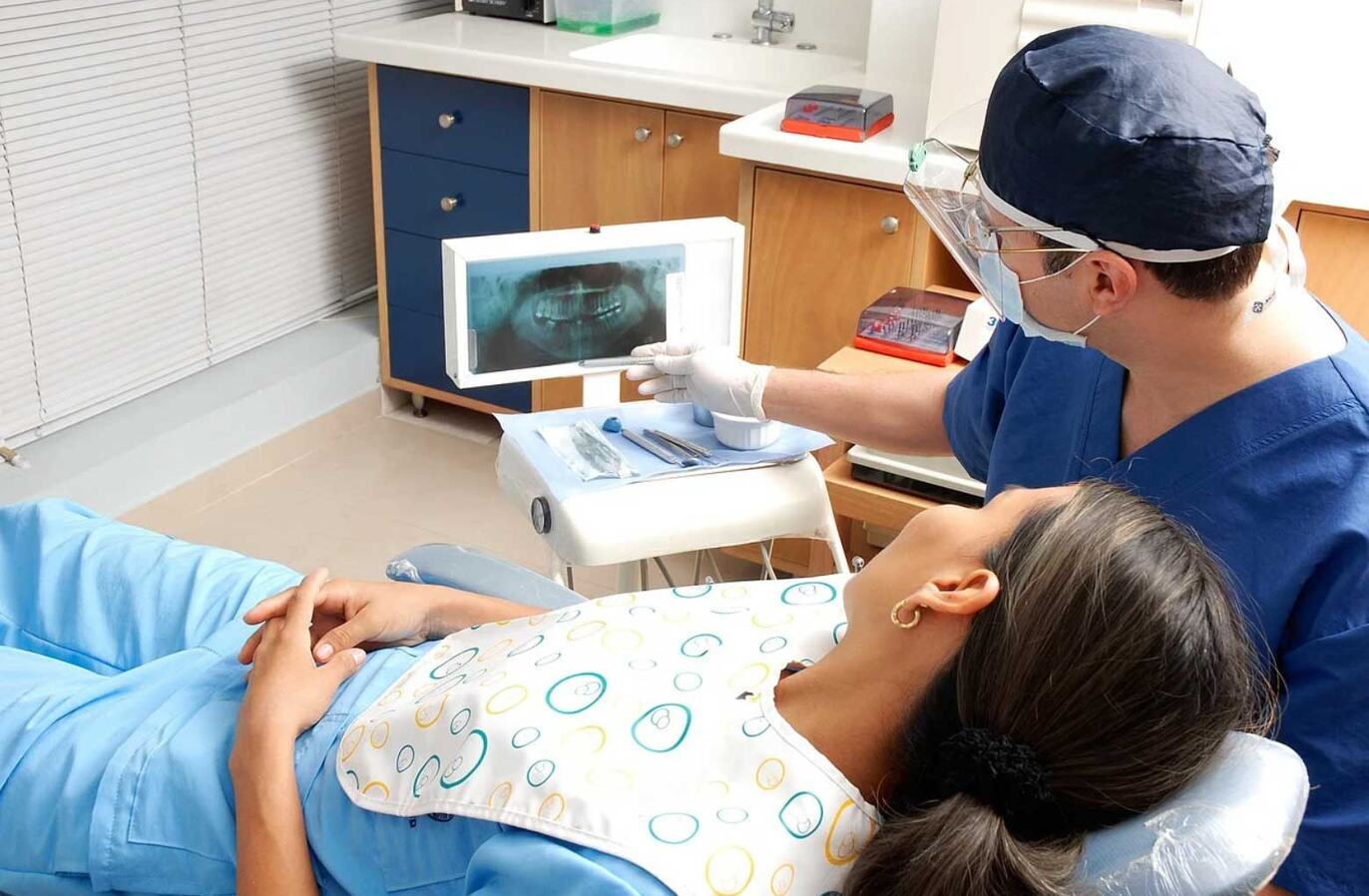 A Los Angeles dentist in blue scrubs, cap, mask and face shield pointing at a patient's panoramic dental X-ray on a light box while the patient reclines in the dental chair.