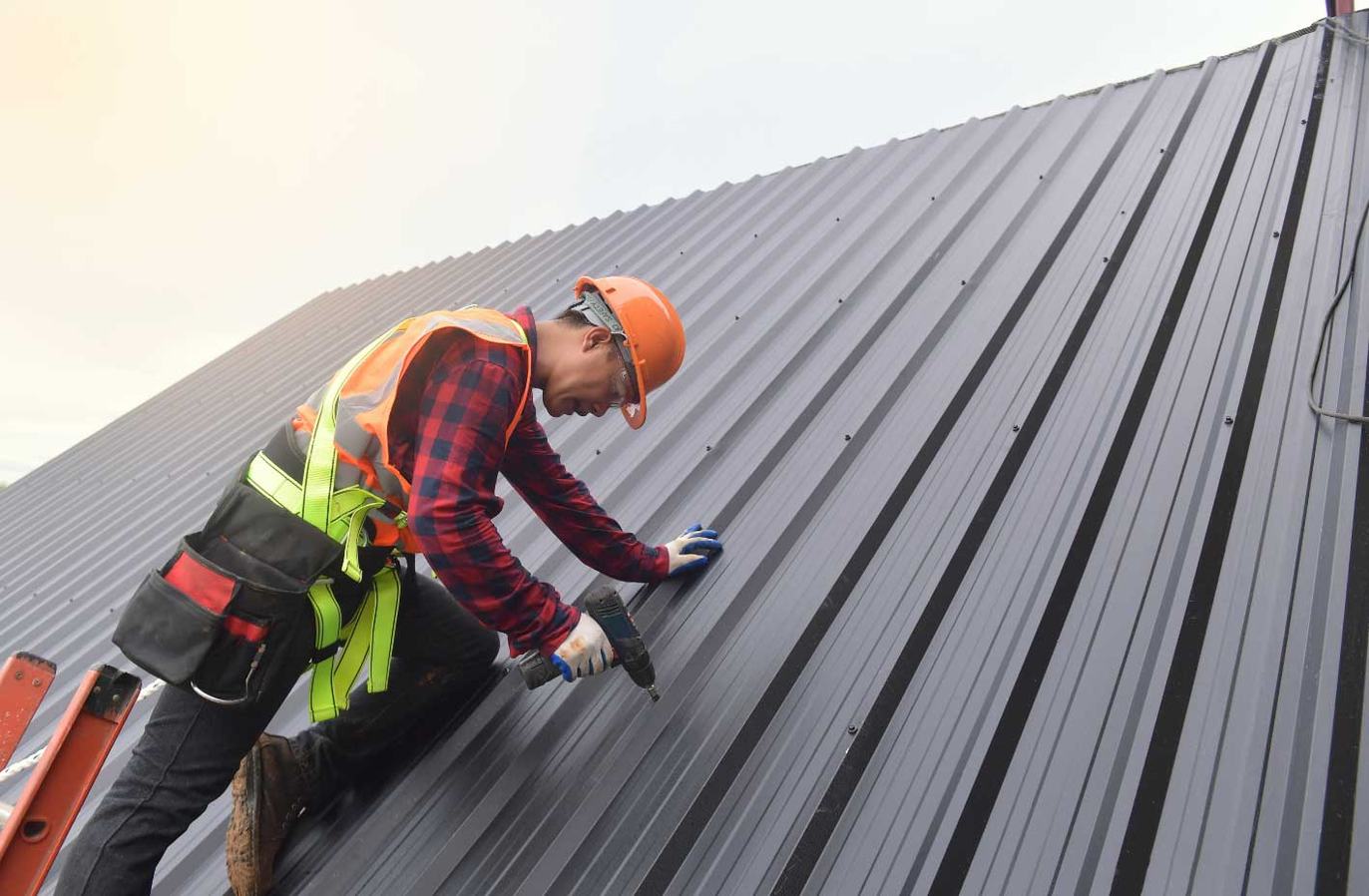 A roofing contractor in a hard hat, safety harness and tool belt kneeling on a dark metal standing-seam roof, driving screws with a cordless drill at dusk.