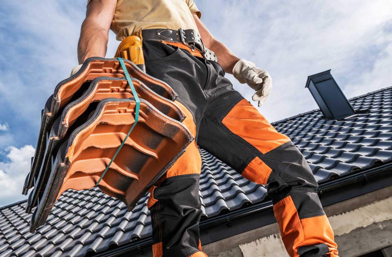A roofing contractor in safety trousers carrying clay roof tiles across a tiled roof, the kind of specialist storm-damage and roof-repair work homeowners search for through AI assistants.