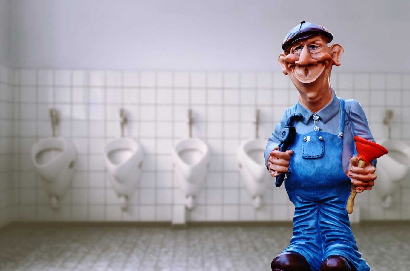 A cartoon plumber figurine in blue overalls and a flat cap holding a wrench and a red plunger, standing in front of a row of white urinals.